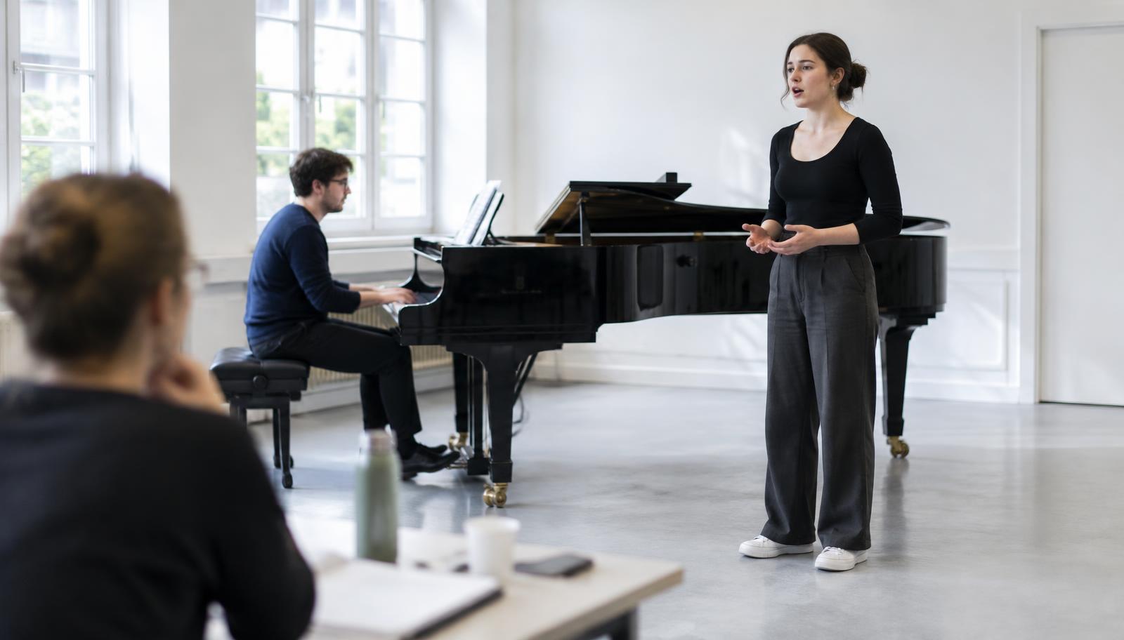 A singer mid-audition beside a grand piano while the accompanist plays, watched from a director's table in a bright rehearsal studio